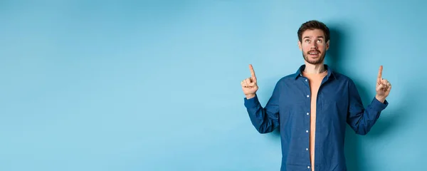 Excited caucasian guy looking and pointing fingers up, showing advertisement, standing on blue background.