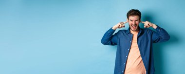 Sinirli genç adam can sıkıcı sesleri engellemek ve kameraya bağırmak için kulaklarını kapatmış. Kaşlarını çatmış ve öfkeli görünüyor. Mavi arka planda duruyor..