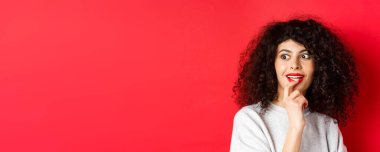 Heyecanlı kıvırcık kadının dudaklarına dokunduğu, ilgiyle yan yana baktığı, havalı tanıtımları yaptığı, kırmızı arka planda dikildiği yakın plan..