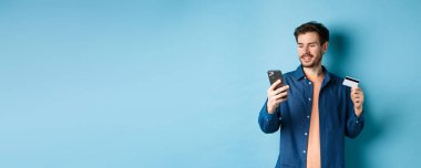 E-ticaret konsepti. Çevrimiçi ödeme yapan yakışıklı beyaz adamın resmi, cep telefonu ekranına bakıyor ve elinde plastik kredi kartı, mavi arka plan.