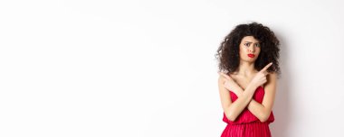 Kırmızı elbiseli, şüpheli ve üzgün genç bir kadın, parmaklarını yana çevirip somurtuyor, ürünler arasında karar veremiyor, seçimlerde yardıma ihtiyacı var, beyaz arka planda duruyor..