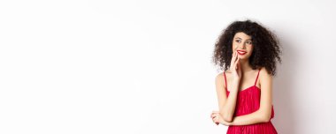 Kıvırcık saçlı, kırmızı elbiseli ve makyajlı, soldaki logoya bakıp gülümseyen, beyaz arka planda duran rüya gibi zarif bir kız..