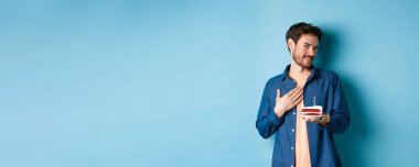 Kutlama ve tatil konsepti. Dokunaklı ve pohpohlanmış bir adam elinde doğum günü pastası ve dokunaklı göğüs minnet, mavi arka planda duruyor..