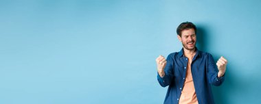 Başarıyı kutlayan mutlu bir adam tasviri, evet diye bağırıyor ve yumruklarını sallıyor, hedefe ulaşıyor ve başarıyor, kazanıyor ve dans ediyor, mavi arka planda duruyor..