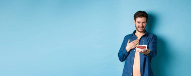 Kutlama ve tatil konsepti. Utanan ve doğum gününü kutlayan adam, yanan mumla doğum günü pastasını uzat, mavi arka planda dur..