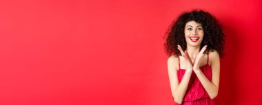 Kıvırcık saçlı, gece elbisesi giyen, el çırpan ve gülümseyen neşeli genç kadın, alkışlarla iyi performansı olan, kırmızı arka planda duran.