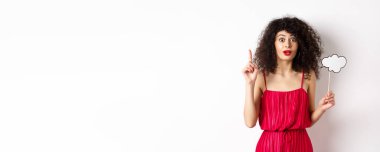 Kırmızı elbiseli hoş beyaz kadın, yorum bulutu tutuyor ve parmağını kaldırıyor, bir fikir ileri sürüyor ve gülümsüyor, bir şeyler öneriyor, beyaz arka planda duruyor..