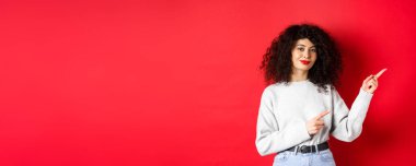 Kıvırcık saçlı, gülümseyen, parmaklarını logoya doğru gösteren, reklam veren, kırmızı arka planlı güzel bir kız..