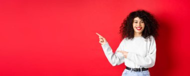 Kıvırcık saçlı, kırmızı dudaklı, gülümseyen beyaz kadın logoyu işaret ediyor, tanıtım teklifini gösteriyor, stüdyo geçmişine karşı duruyor..