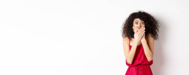 Şok olmuş bir kadın elleriyle ağzını kapatıyor ve kameraya güvensizlikle bakıyor. Şok edici bir şeye tanık oluyor. Beyaz arka planda kırmızı elbiseyle dikiliyor..