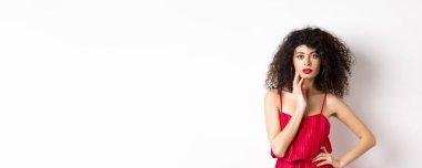 Güzellik ve moda. Şehvetli, kıvırcık saçlı, kırmızı dudaklı, zarif bir elbise giyen, yüzüne dokunan ve baştan çıkarıcı görünen beyaz bir kadın model..