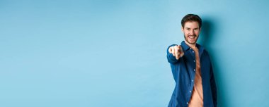 Kendinden emin gülümseyen adam seni seçiyor ya da davet ediyor, kameraya işaret ediyor kararlı, mavi arka planda duruyor. Boşluğu kopyala