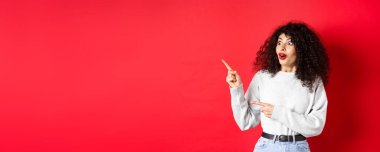 Şaşırmış kıvırcık saçlı, boş alanı gösteren, logo ya da reklam veren, kırmızı arka planda duran, kıvırcık saçlı, çekici kız..
