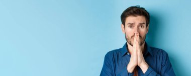 Üzgün genç adama yaklaş, yardım için yalvar ve lütfen de, mavi arka planda dur..