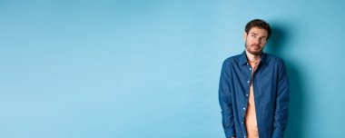 Gönülsüz ve üzgün bir adam üzgün hissediyor, boş bir yere bakıyor, güler yüzlü, mavi arka planda duruyor..