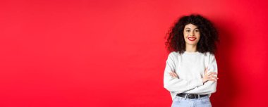 Makyajlı, kıvırcık saçlı, göğsünde çapraz kolları olan ve profesyonel görünümlü, kırmızı arka planı olan kendine güvenen bir kadın..
