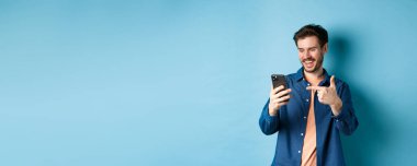 Mobil ekranı okuyan, telefonu işaret eden, online tanıtım ya da akıllı telefon uygulamasını kontrol eden, mavi arka planda duran yakışıklı bir adam resmi.