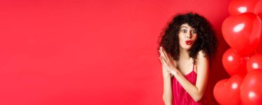 Heyecanlı kıvırcık saçlı güzel bir kadın, kalp balonlarının yanında duruyor ve avuç içlerini birbirine sürtüyor, iyi bir anlaşma ya da zevk bekliyor, kırmızı arka plan.