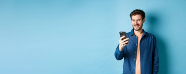Çekici beyaz adam cep telefonu kullanıyor, mutlu görünüyor ve kameraya gülümsüyor, mavi arka planda duruyor..