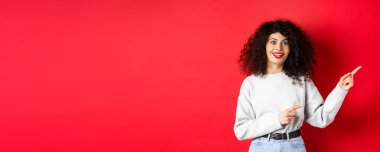Kıvırcık saçlı neşeli genç kadın, parmaklarını logoya doğrultuyor, etkilenmiş ve heyecanlı görünüyor, terfiyi kontrol ediyor, kırmızı arka planda..
