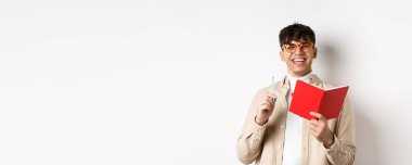 Gözlüklü neşeli genç adam gülüyor ve not alıyor, planlayıcıyla yazıyor, elinde kalem ve günlükle beyaz arka planda duruyor..