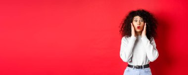 Şaşırmış genç bir kadın inanılmaz haberler duyuyor, tanıtıma bakıyor ve diyor ki, vay canına, inançsızlıkla bak, kırmızı arka planda dur..