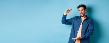 Neşeli modern adam gülümsüyor, büyük boy gösteriyor ve mutlu görünüyor, mavi arka planda duruyor. Boşluğu kopyala