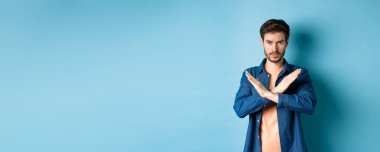 Ciddi beyaz adam hayır diyor, çapraz duruş hareketi yapıyor ve kameraya kendinden emin bakıyor, kötü bir şeyi yasaklıyor, mavi arka planda duruyor..