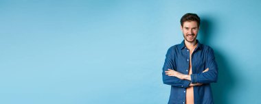 Kendine güveni tam genç bir adam kendini profesyonel hissediyor, kollarını göğsünde kavuşturuyor ve sırıtıyor, mavi arka planda duruyor..