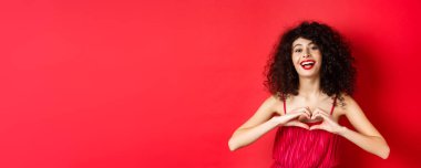 Kıvırcık saçlı, kırmızı elbiseli, kalp sembolü gösteren ve gülümseyen mutlu, stüdyo geçmişi olan güzel bir kadın..