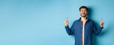 Şüpheci genç adam sırıtıyor ve şüpheli bakıyor, üzgün parmaklarını gösteriyor, mavi arka planda duruyor. Boşluğu kopyala