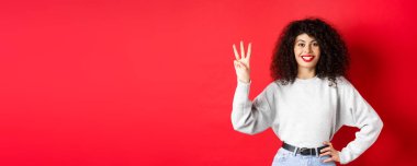 Üç numarayı gösteren, gülümseyen, sipariş veren, kırmızı arka planda eşofman üstü giyen neşeli bir kadın model..