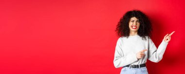 Kuşkucu Kafkasyalı kadın suratını buruşturuyor, zevksizlikten şikayet ediyor, parmaklarını sağa gösteriyor ve hoşlanmamaktan çekiniyor, kırmızı arka plana karşı duruyor..