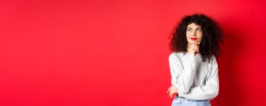 Dalgın, kıvırcık saçlı, kırmızı dudaklı, sol üst köşedeki logoda düşünceli görünen, gülümseyen, stüdyo arka planında duran....