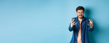 Öfkeli adam birini boğmak ister, düşmanını boğmak için ellerini uzatır, dudaklarını ısırır ve öfkeli görünür, mavi arka planda durur..