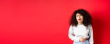 Kadın hasta, eğiliyor ve karnına dokunuyor, karın ağrısı ya da regl sancısı çekiyor, kırmızı arka planda duruyor..