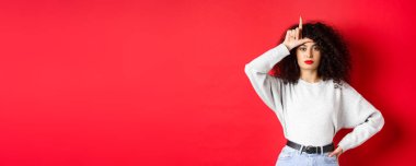 Kıvırcık saçlı, kendine güveni tam bir Avrupalı kadın, kayıp takımla alay ediyor, ezik jestleri yapıyor ve kameraya bakmıyor, kırmızı arka planda sıradan kıyafetler giyiyor..