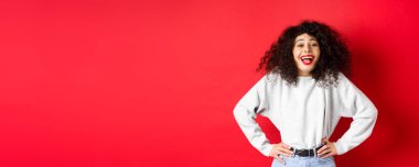 Heyecanlı pozitif kadın gülüyor ve gülümsüyor, neşeli hissediyor, kırmızı arka planda duran komik tanıtıma bakıyor..