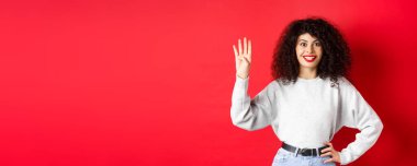 Kıvırcık saçlı heyecanlı kadın parmaklarıyla dört numarayı gösteriyor, düzen kuruyor, kırmızı arka planda duruyor..