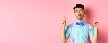 Papyonlu neşeli komik adam parmaklarını kaldırıp reklamını yapıyor ve kararsız gülümsüyor pembe arka planda dikiliyor. Boşluğu kopyala