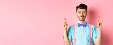 Umut dolu genç bir adam dilek diliyor, parmaklarını çapraz yapıp gülümsüyor, iyimserlikle sonuçları bekliyor, pembe arka planda duruyor..