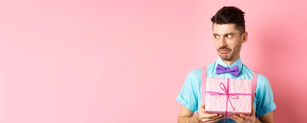 Holidays and celebration concept. Shocked guy staring aside with cringe face, holding small gift box, standing over pink background.