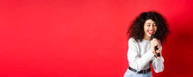 Hobiler ve eğlence konsepti. Güzel bir kadın mikrofonda şarkı söylüyor ve gülümsüyor, karaoke 'de eğleniyor, kırmızı arka planda duruyor..