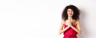 Kırmızı dudaklı çekici bir genç kadın, gece elbisesi, teşekkür ediyor ve gülümsüyor, namaste jesti ile minnettar görünüyor, beyaz arka planda duruyor..