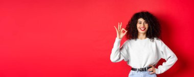 Kendine güvenen güler yüzlü kadın, onaylamak ya da ürünleri sevmek için onay işareti gösteriyor, iyi kalite garanti ediyor, iltifat ediyor, kırmızı arka plan.