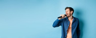 Kaygısız adam mikrofonla karaoke yapıyor, şarkı söylüyor, mavi arka planda duruyor. Boşluğu kopyala