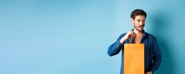 Turuncu alışveriş torbası gösteren, göz kırpan, mağaza indirimi öneren, mavi arka planda duran mutlu genç adam..
