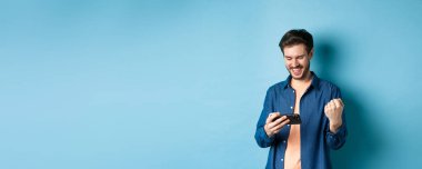 Cep telefonuyla video oyununda kazanan mutlu genç adam, akıllı telefon ekranına bakıyor ve sevinçle evet diyor, mavi arka planda duruyor..