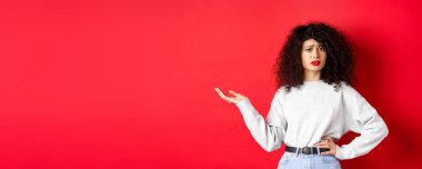 Kafası karışmış ve şüpheci bir kadın, kıvırcık saçlı, kaşlarını çatan ve el kaldıran, kırmızı arka planda duran garip bir şeyi anlayamıyor..