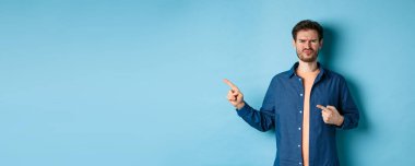 Hüsrana uğramış ve şüpheci adam somurtuyor, dudaklarını büzüyor ve parmaklarını boş yere doğrultuyor, kötü şeyden yakınıyor, mavi arka planda üzgün duruyor..